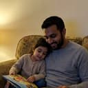 A dad on the sofa reading a picture book with his young daughter leaning into his side.