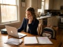 A mum on the phone at her kitchen table, laptop open, family planner and school letters in front of her.
