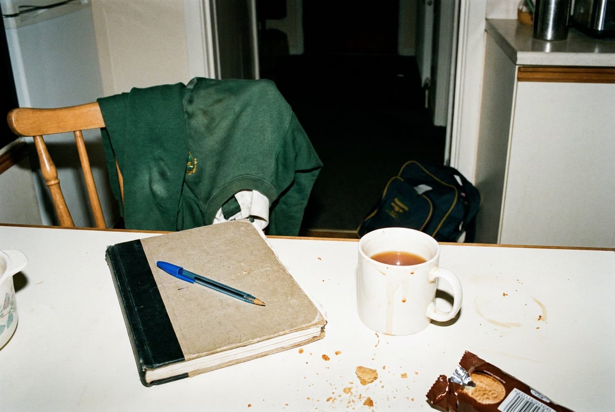 A kitchen table at night lit by harsh camera flash: a closed notebook with a biro on top, a half-drunk mug of tea, a school jumper over the chair and a school bag dumped in the dark hallway behind