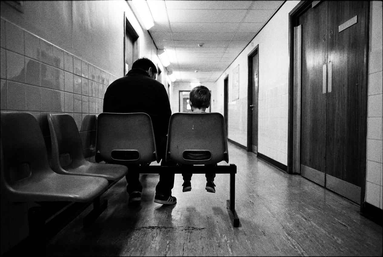 A parent and young child seen from behind, sitting together on chairs in an empty hospital outpatient corridor