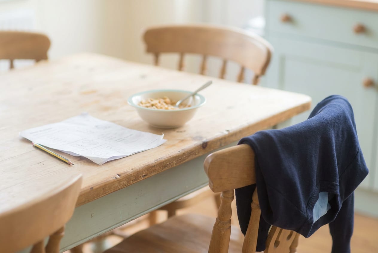 A kitchen table in pale morning light, with last night's unfinished homework sheet and pencil beside an untouched bowl of cereal and a school jumper over a chair.