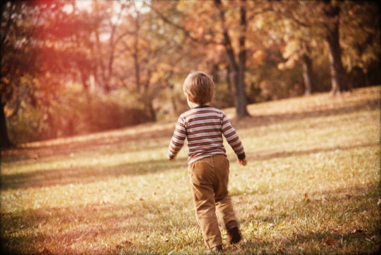 A faded film snapshot of a park on a Saturday, a small child in a coat running far ahead across the grass, seen from behind at a parent's distance.