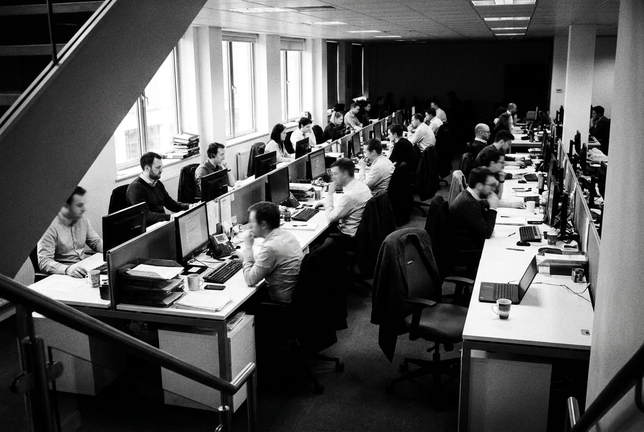 A wide black-and-white view across a busy open-plan office floor, rows of desks with people working, every face turned away or blurred, and one empty chair with a coat still over the back