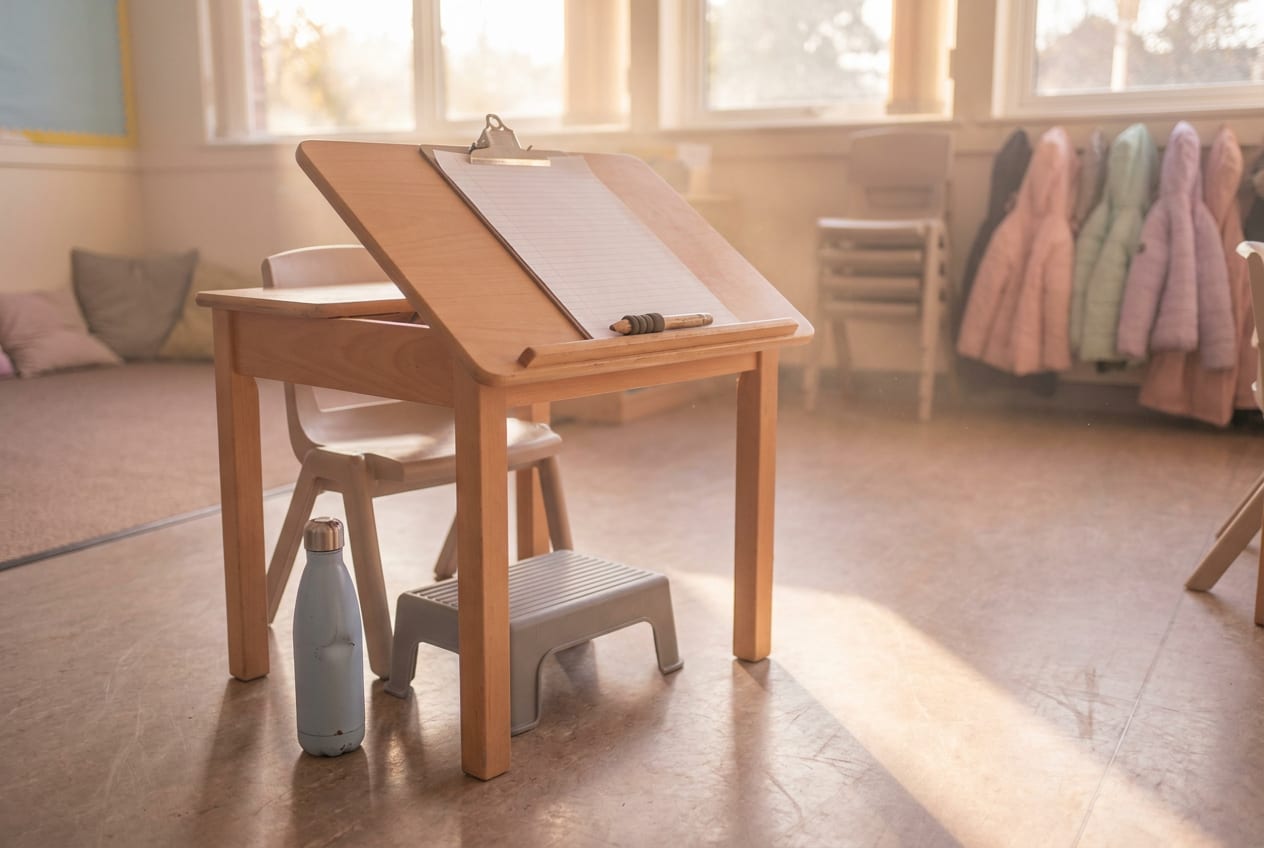 An empty primary school desk at the end of the day, set up with a sloped writing board, a pencil with a moulded grip, and a small footrest under the chair.