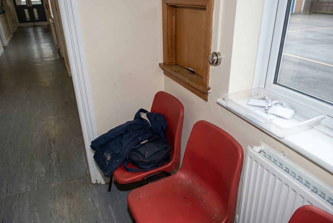 Two low chairs in an empty school reception waiting area on a weekday morning, with a child's coat and book bag left on one of them and a closed office hatch behind.
