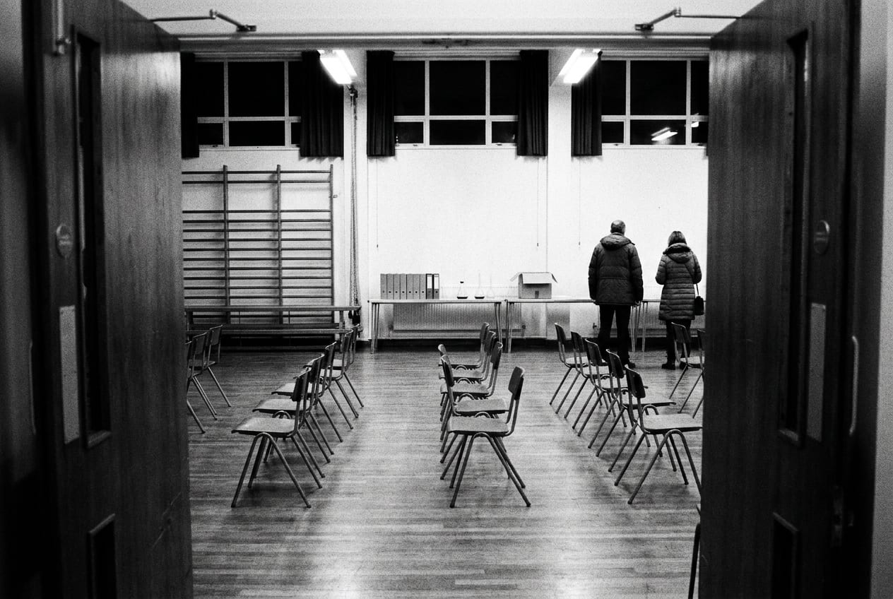 A secondary school hall set out for an open evening on an autumn night, stacking chairs pulled out of line and a trestle table along one wall, the strip lights on against dark windows.
