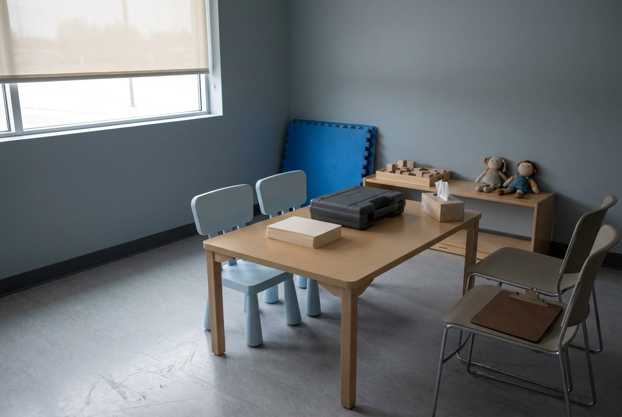 An empty clinic assessment room set up before an appointment: a low table with small chairs on one side and adult chairs on the other, blank picture cards, a closed case of assessment materials, and simple toys on a shelf.