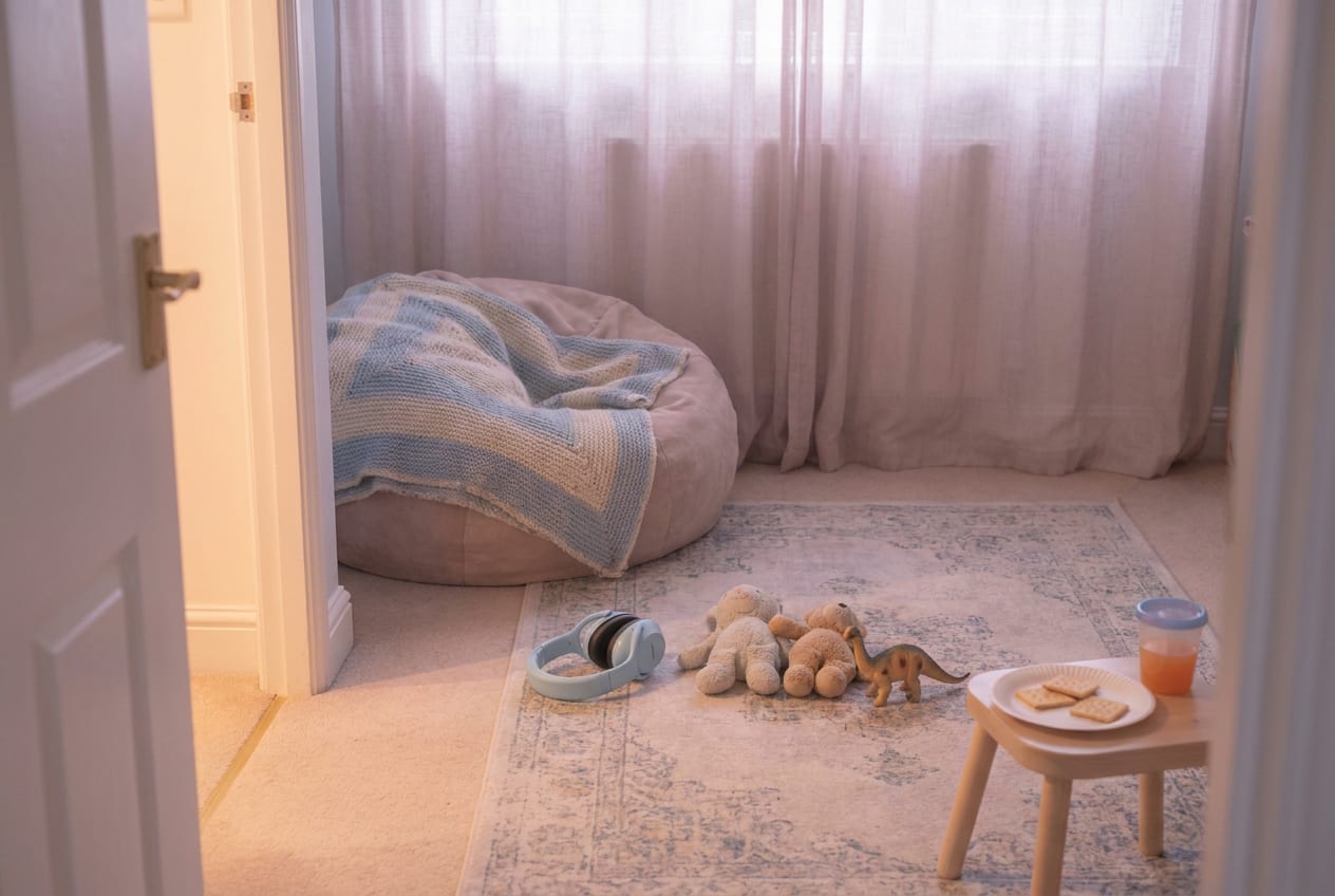 A small quiet bedroom set up during a birthday party downstairs: a beanbag with a familiar blanket, child-sized ear defenders on the carpet, two soft toys and a plate of plain crackers on a low stool, soft light through thin curtains