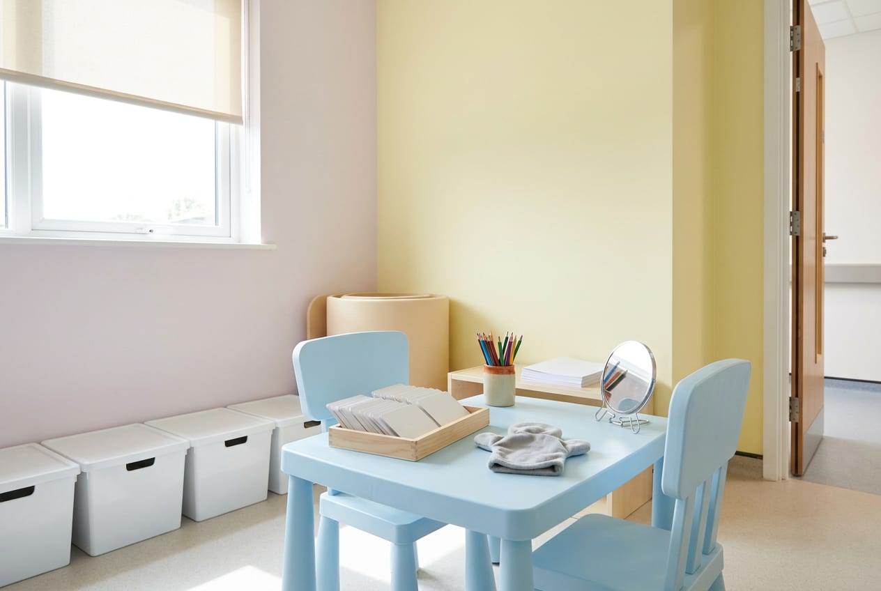 An empty speech and language therapy room, brightly lit, with two small chairs at a low table and a tray of picture cards set out ready.