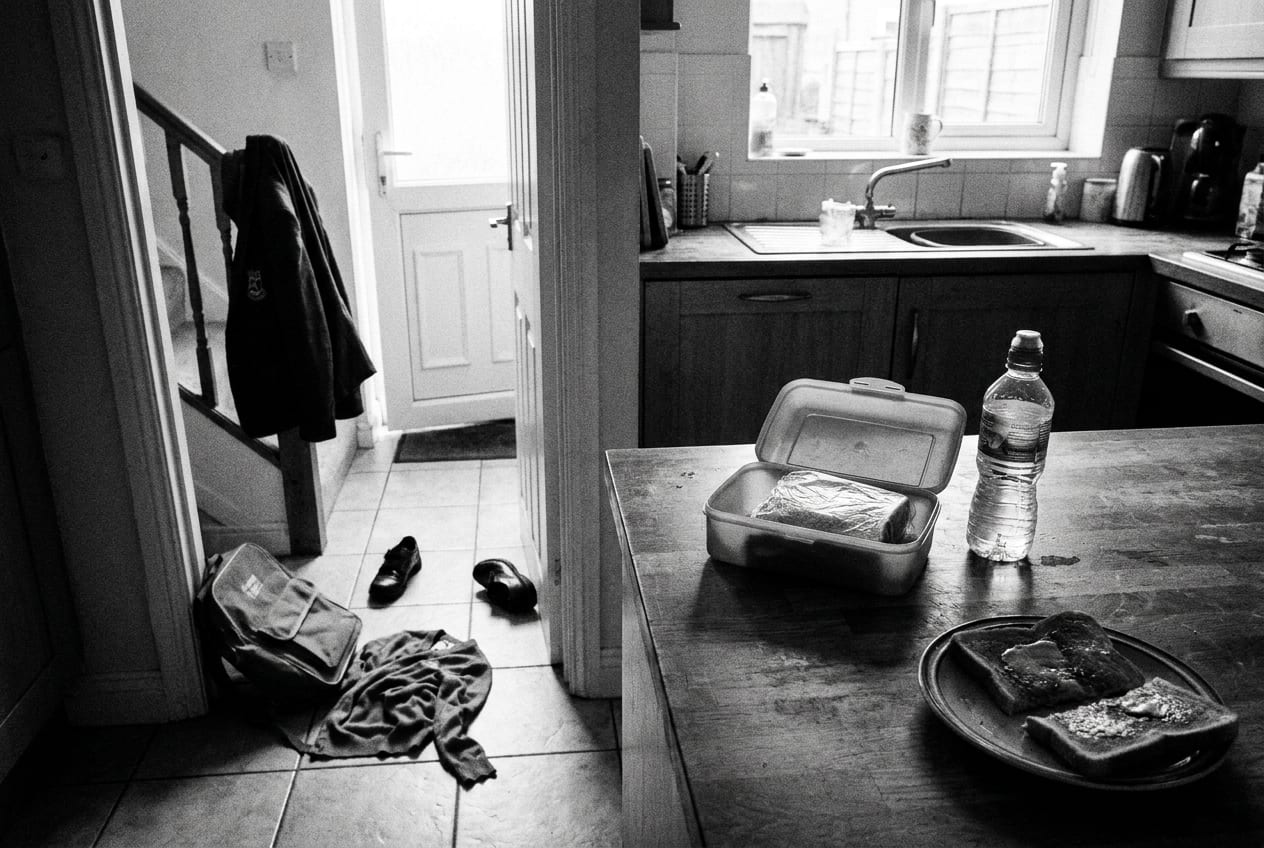 A kitchen worktop in the afternoon with an open lunchbox holding an untouched wrapped sandwich and a full water bottle, a half eaten slice of toast on a plate, a school bag dropped on the floor and shoes kicked off in the hallway beyond