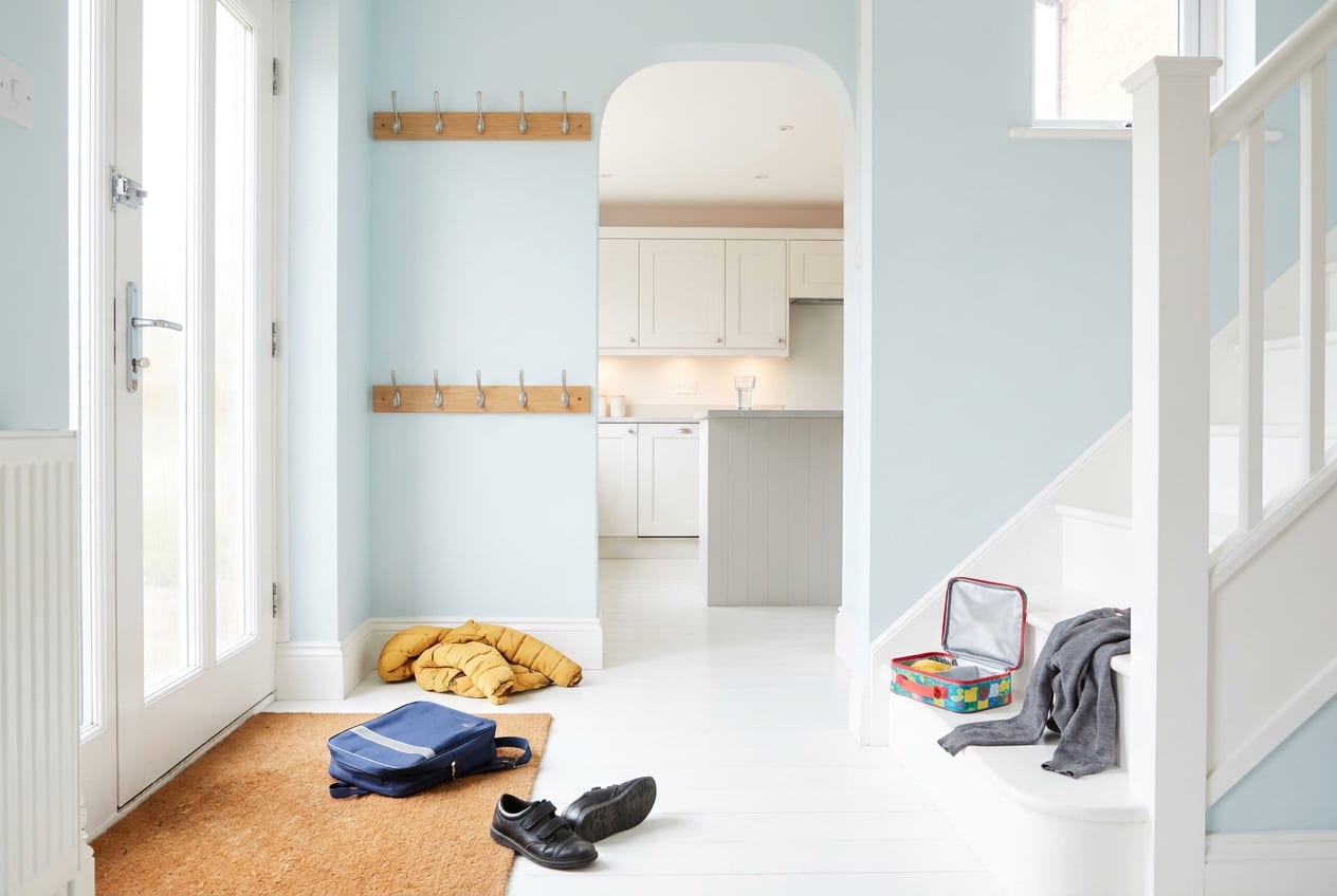 An empty, brightly lit hallway inside a British family home in the afternoon, with a school book bag dropped on the floor, shoes kicked off on the mat and a coat fallen beneath its hook.