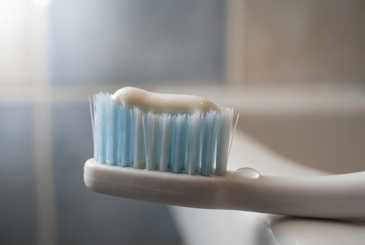 Extreme close-up of a small toothbrush with a stripe of toothpaste already squeezed onto the bristles, resting on a bathroom shelf, most of the frame falling out of focus